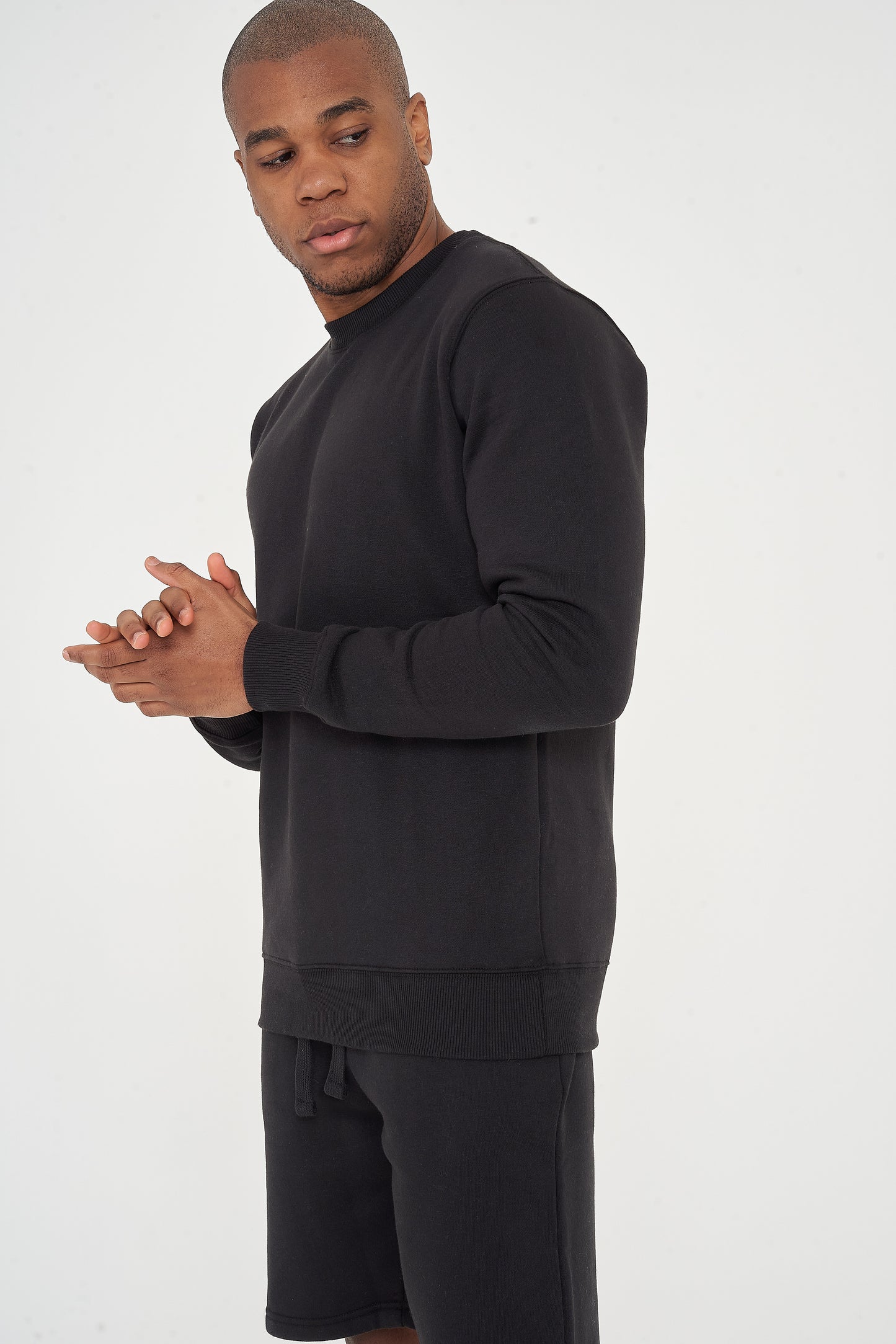 Male model side view wearing black 365 Regular Sweatshirt and shorts, hands clasped – regular fit, 365 Collection.