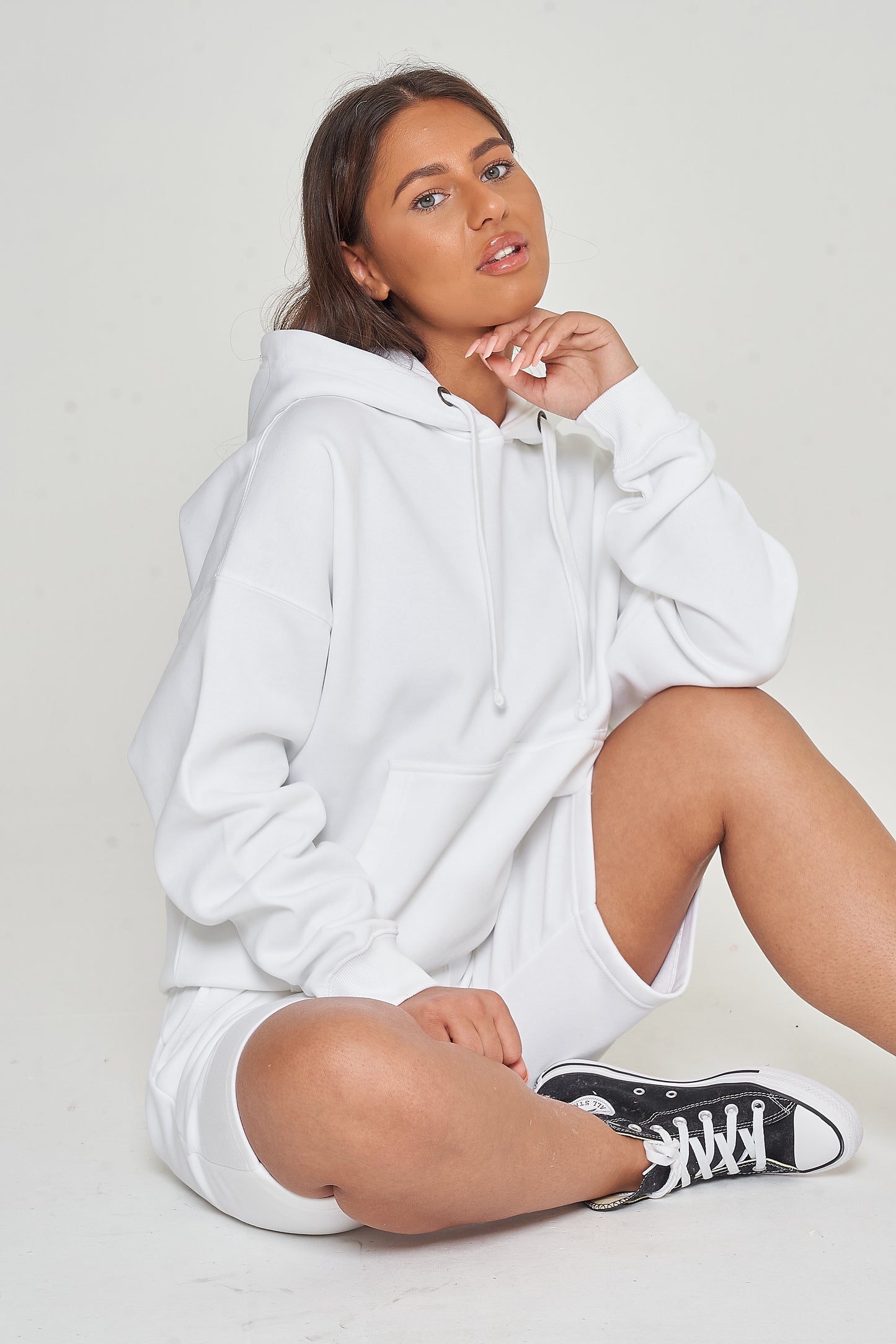 A woman sitting in an oversized white hoodie and shorts, showing a relaxed streetwear style.