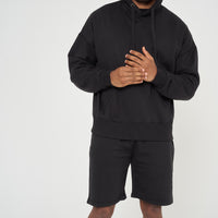 Male model side angle wearing black 365 Oversized Hoodie with hood up and shorts – oversized fit, 365 Collection.