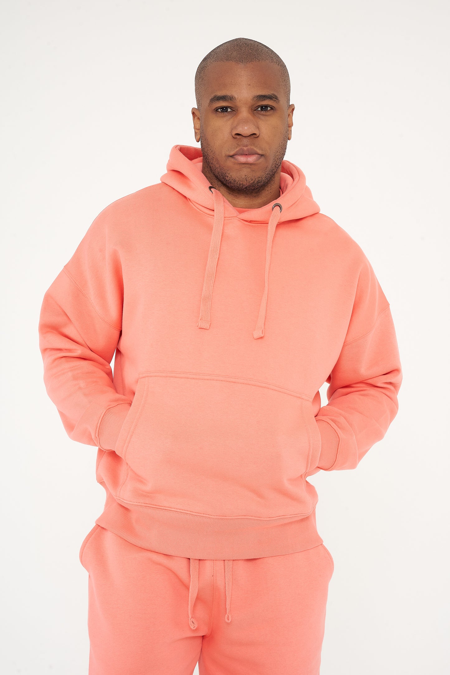 Male model standing in coral 365 Oversized Hoodie and shorts with hands in pockets, relaxed posture – 365 Collection – oversized fit. 
