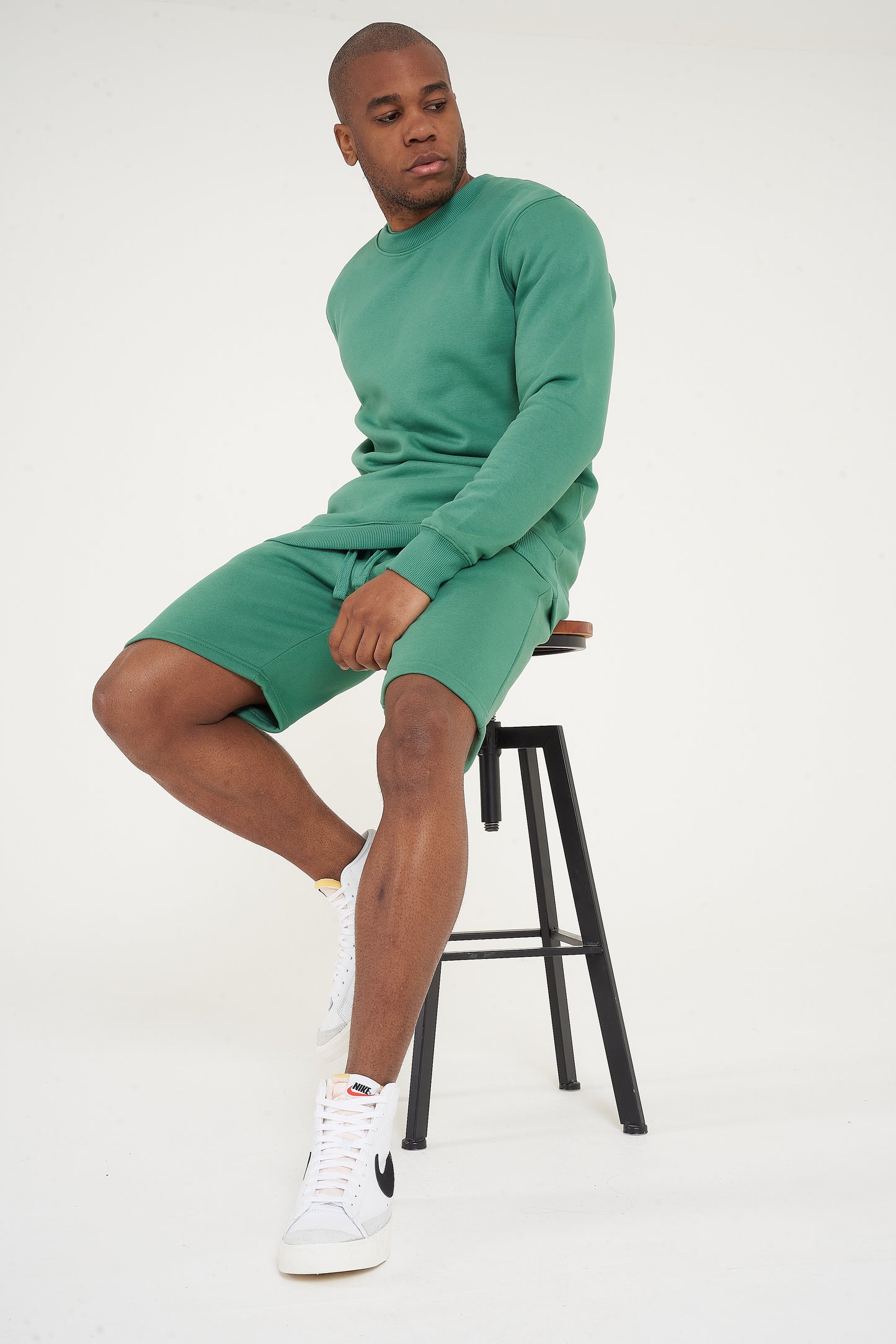 Male model seated wearing green regular fit sweatshirt and shorts, casual pose - 365 Collection.
