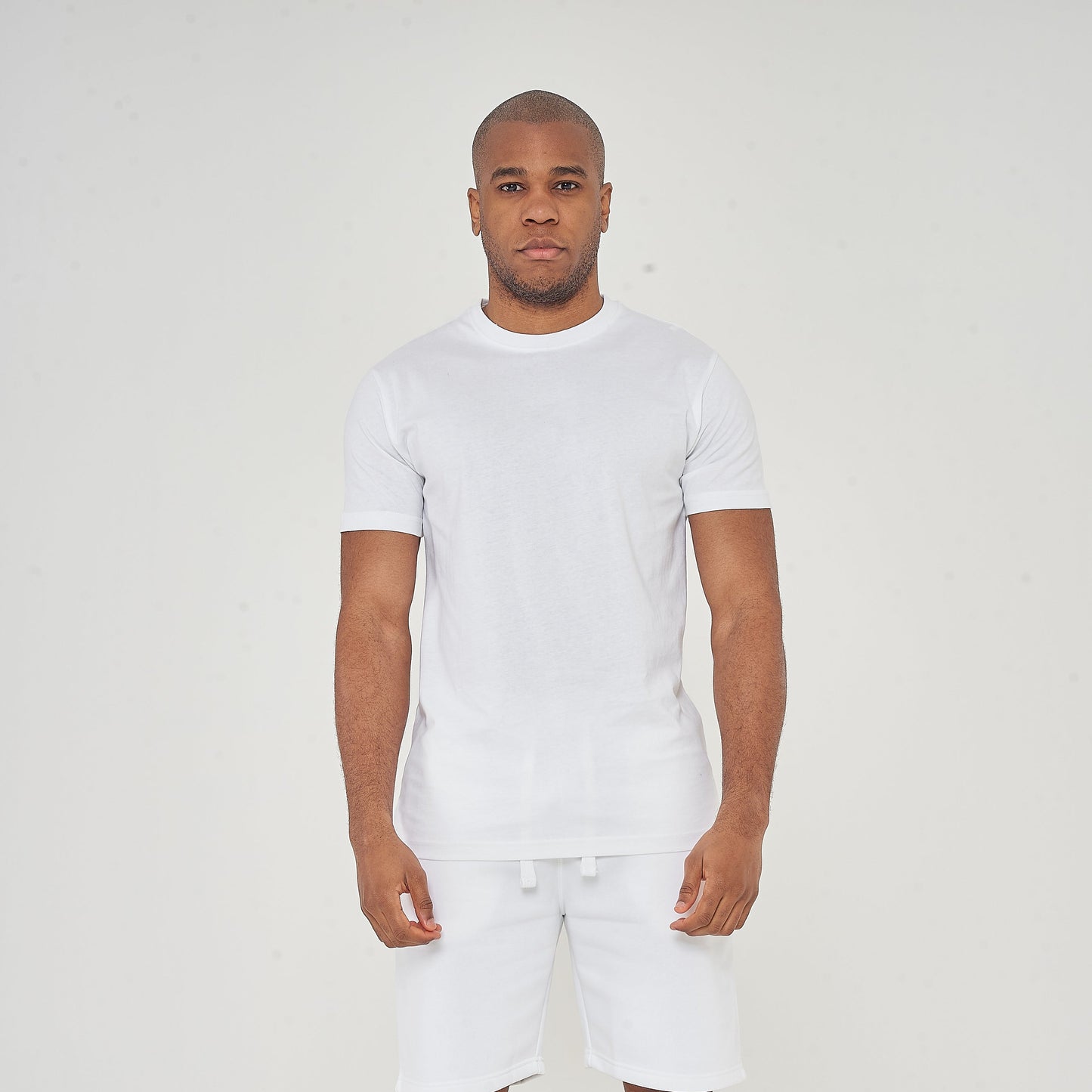 Man standing in white t-shirt and white shorts with black sneakers on a plain light background.