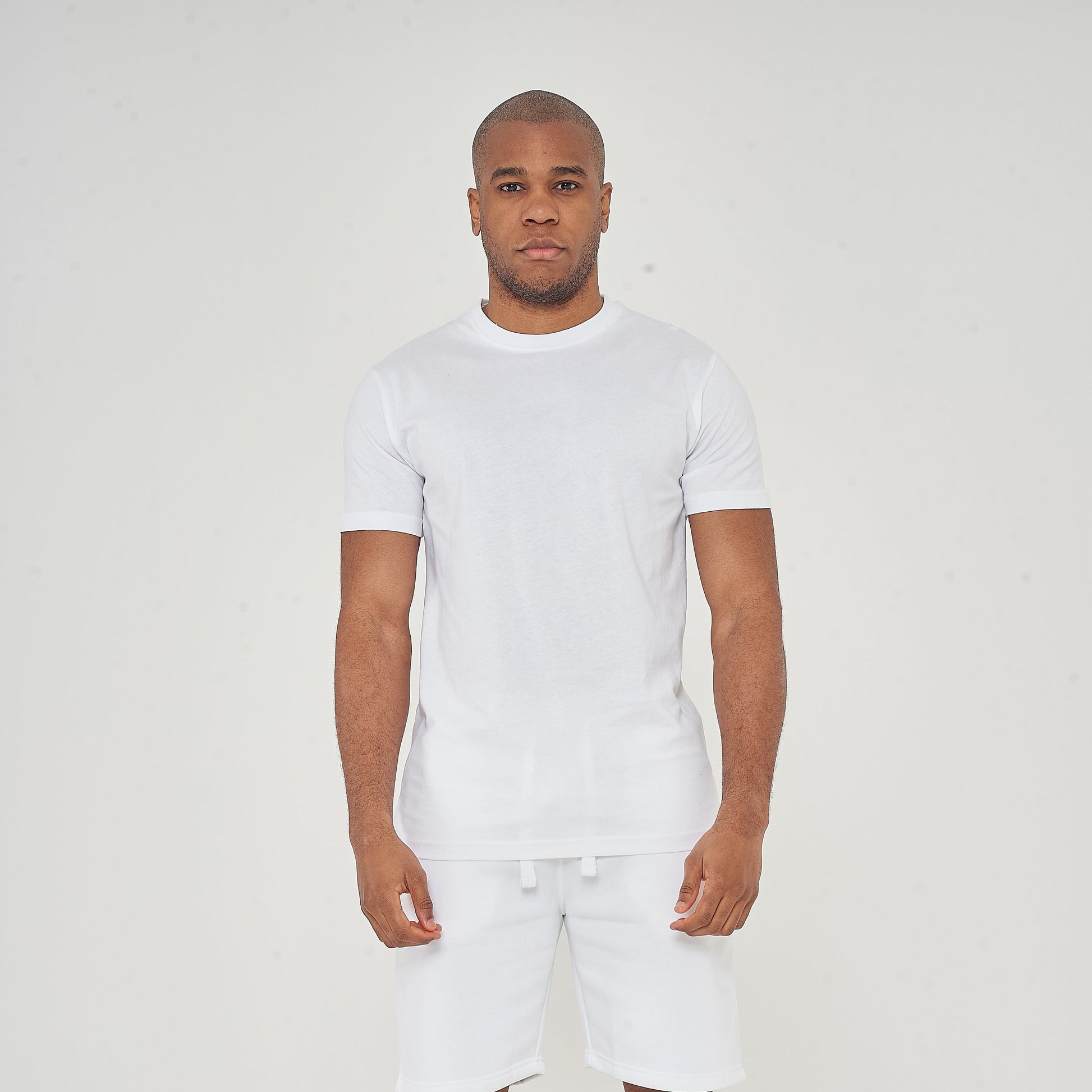 Man standing in white t-shirt and white shorts with black sneakers on a plain light background.