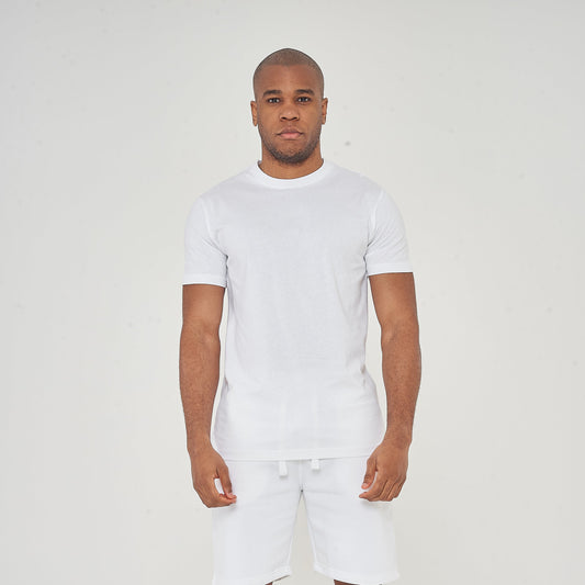 Man standing in white t-shirt and white shorts with black sneakers on a plain light background.