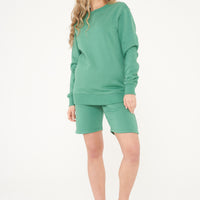 A woman wearing a basic green sweatshirt and matching shorts, standing front-facing. 