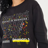 Woman wearing the black New York Season 1 Sweatshirt styled with casual bottoms, highlighting the prominent front graphic and comfortable oversized fit.