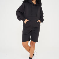 A woman wearing an oversized black hoodie and shorts, standing front-facing, showing a loose fit. 