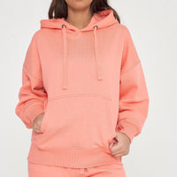 Woman modeling oversized coral hoodie and shorts, standing with hands in front pocket. 