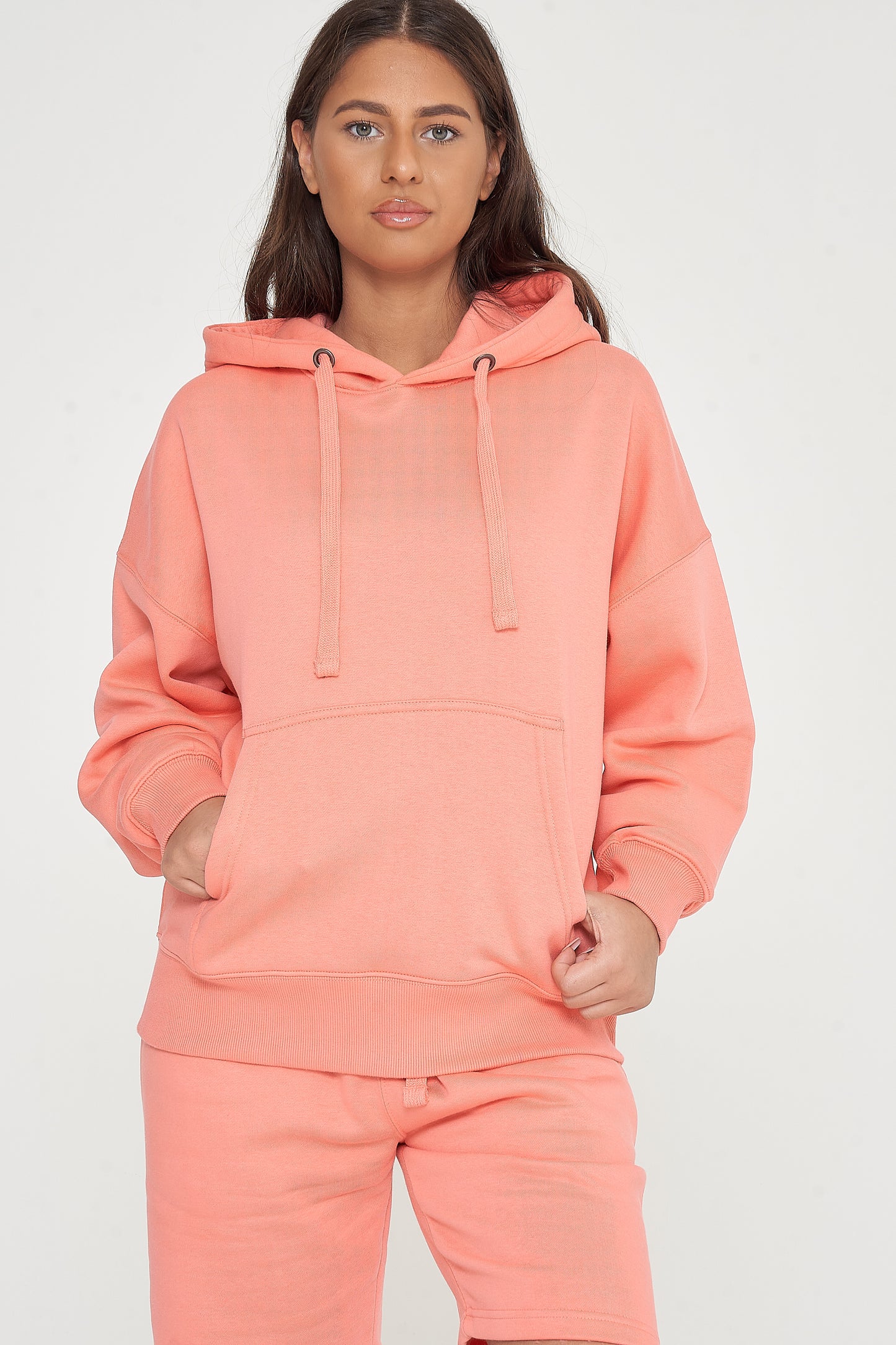Woman modeling oversized coral hoodie and shorts, standing with hands in front pocket. 