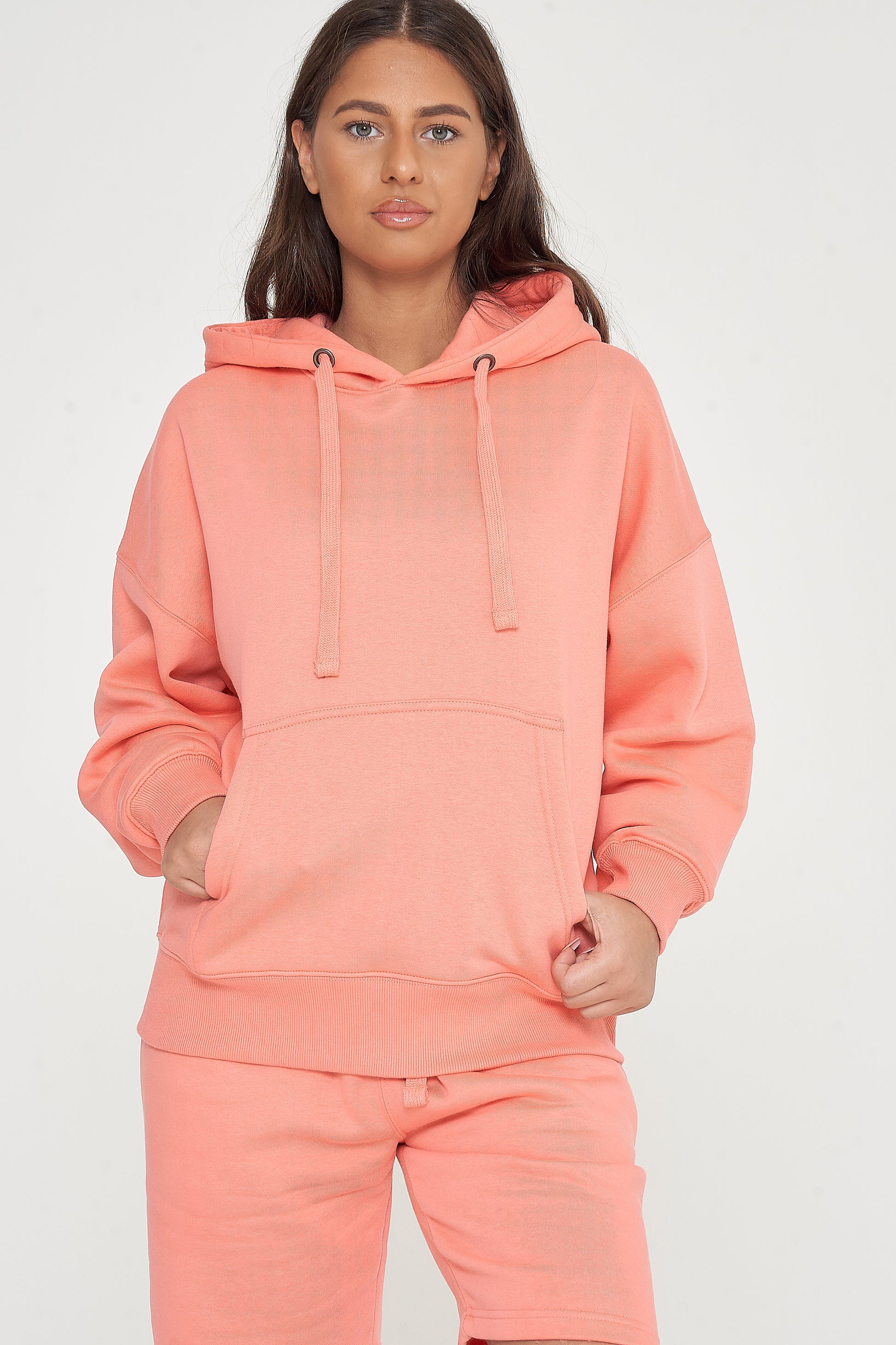 Woman modeling oversized coral hoodie and shorts, standing with hands in front pocket. 