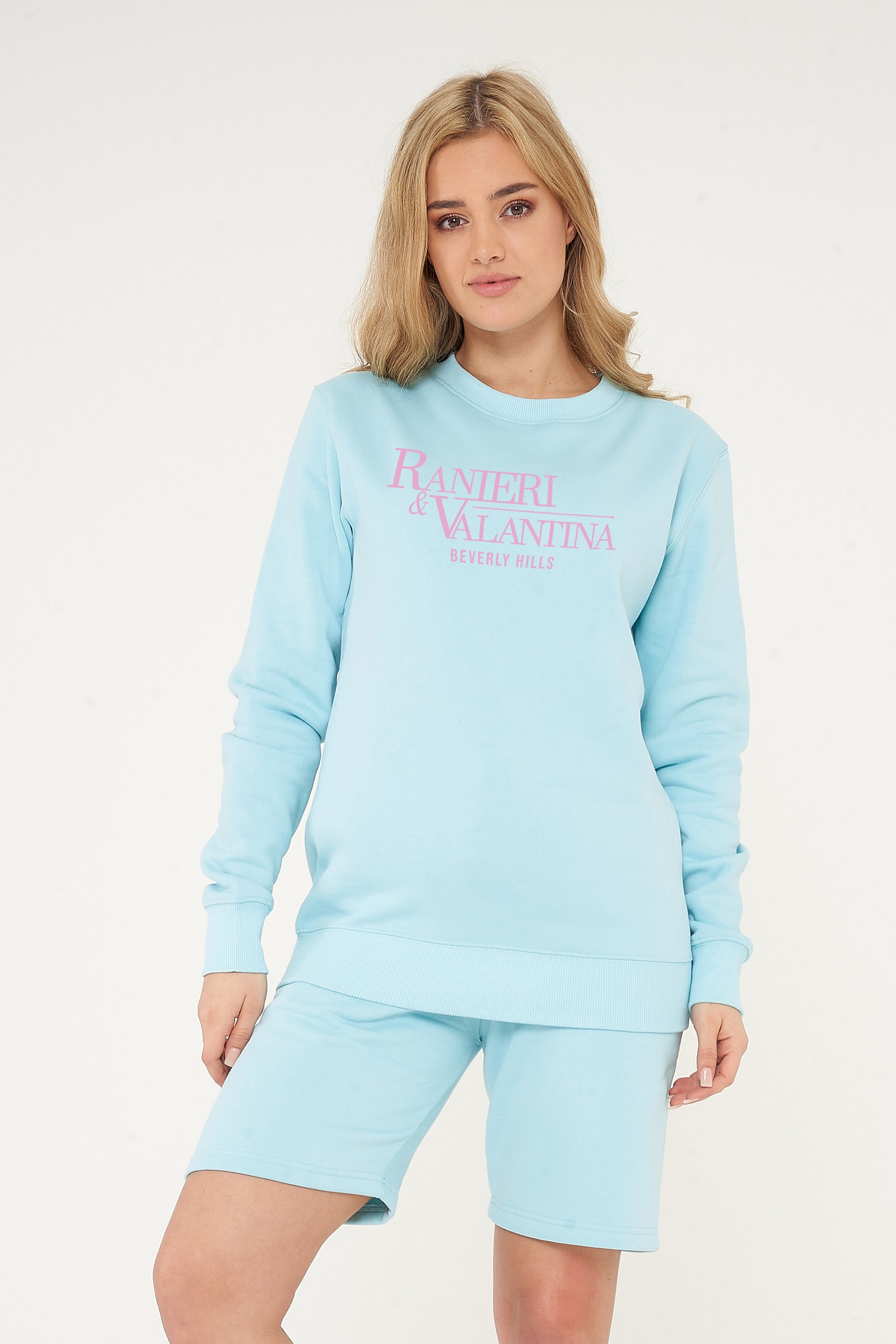 Woman wearing turquoise Signature Beverly Hills sweatshirt styled with matching shorts, standing against neutral background.